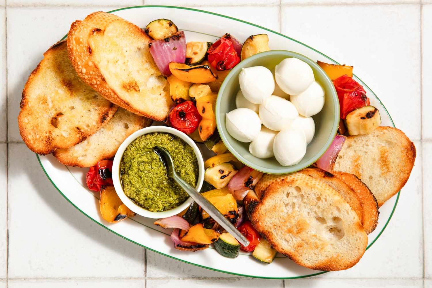 A plate with toasted bread, fresh mozzarella, grilled vegetables, and a bowl of pesto