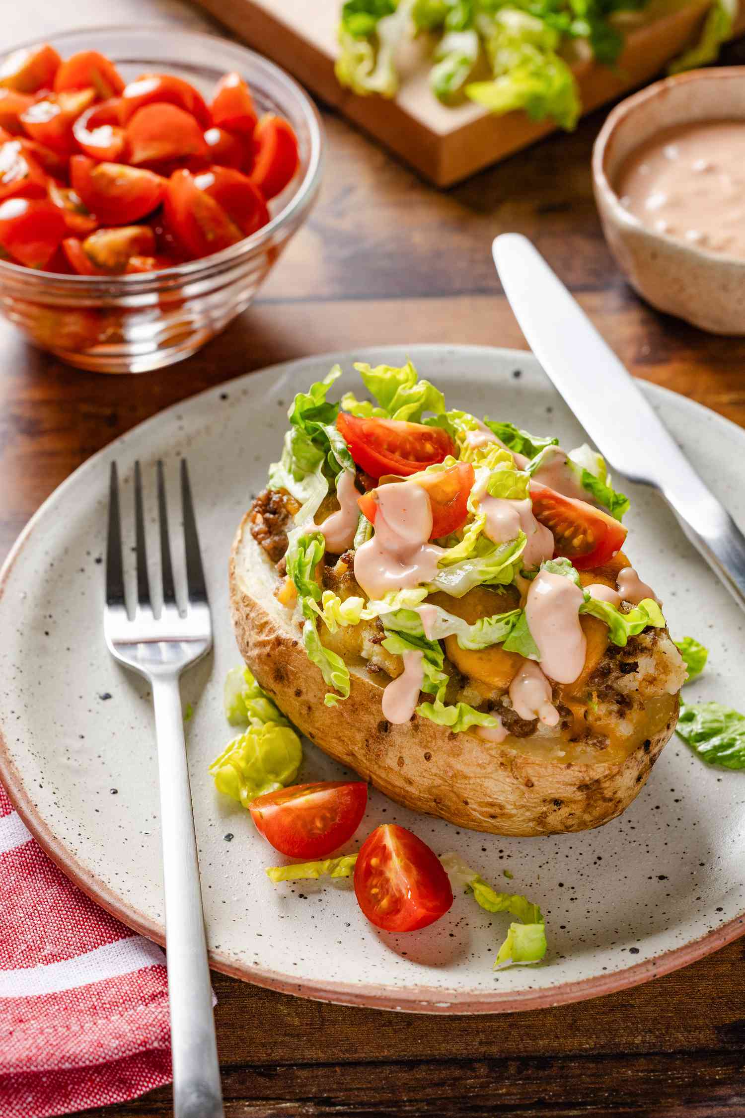 Cheeseburger stuffed baked potatoes with toppings on a plate with utensils and in the surroundings, a bowl of sliced tomatoes, bowl of sauce, and chopped lettuce