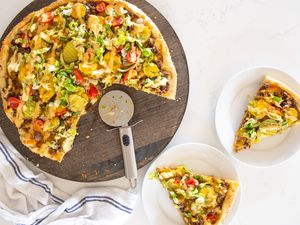 Slice of Cheeseburger Pizza on Small Plate Next to a Circular Wooden Tray with More Pizza and a Pizza Wheel and on the Counter, a Kitchen Towel