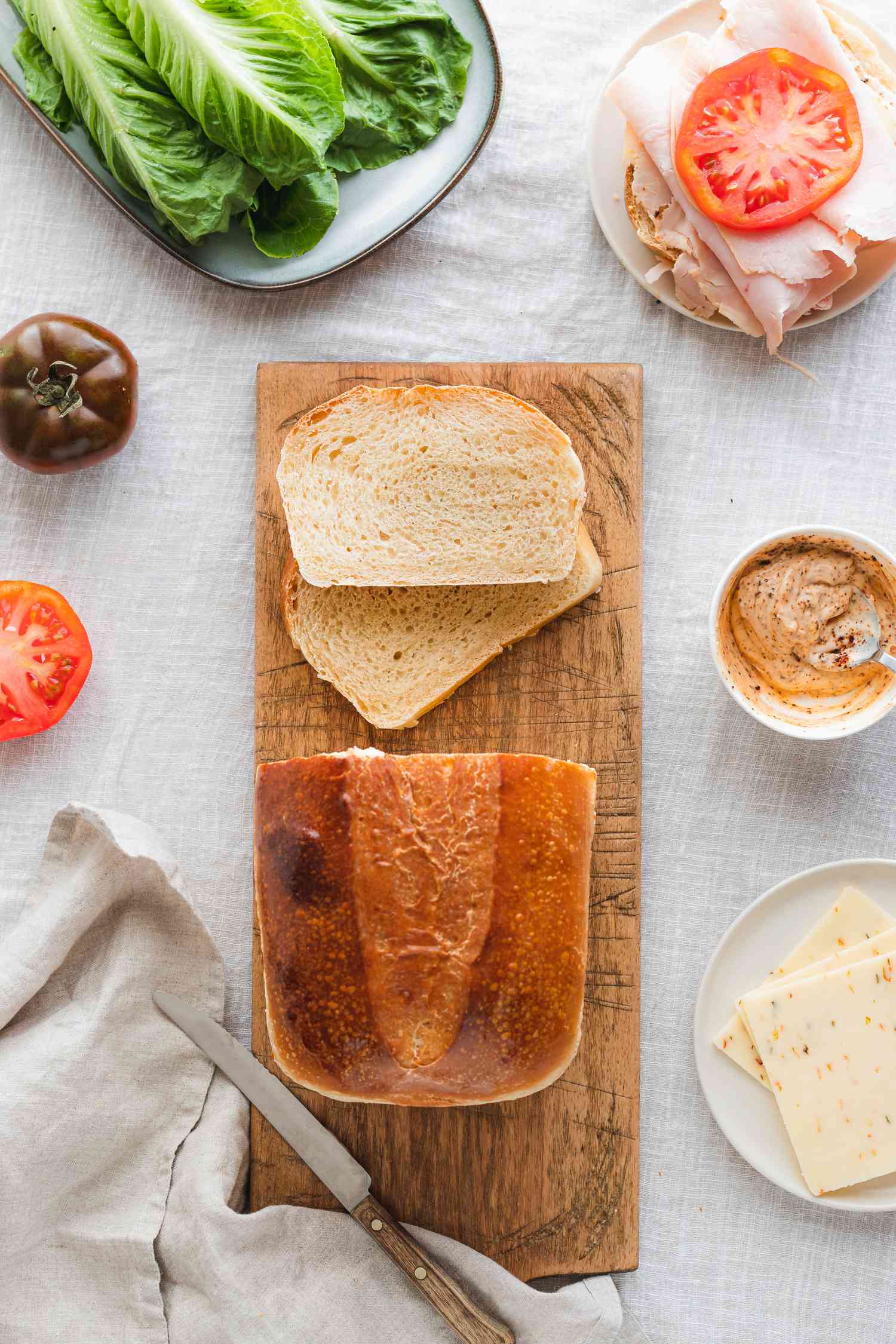 Sourdough Sandwich Loaf with Slices Cut on a Cutting Board, Surrounded by Sandwich Ingredients (a Small Plate with Cheese, a Platter with Lettuce, a Small Plate with a Slice of Bread Topped with Turkey Slices and Tomato Slice, a Small Bowl with a Sandwich Sauce, and a Sliced Tomato)