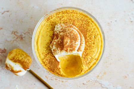 Overhead shot: 2-ingredient pumpkin mousse topped with whipped cream and a sprinkle of pumpkin spice, all in a sherbert glass. On the counter, a spoonful of mousse