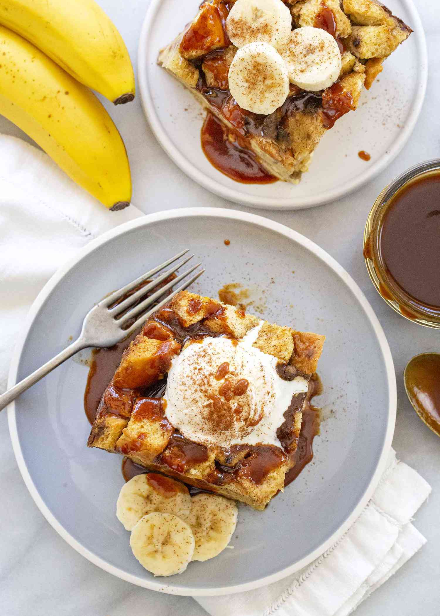 Slice of the best slow cooker banana bread pudding topped with a scoop of ice cream and drizzeld with caramel sauce on a gray plate with a fork and sliced bananas.