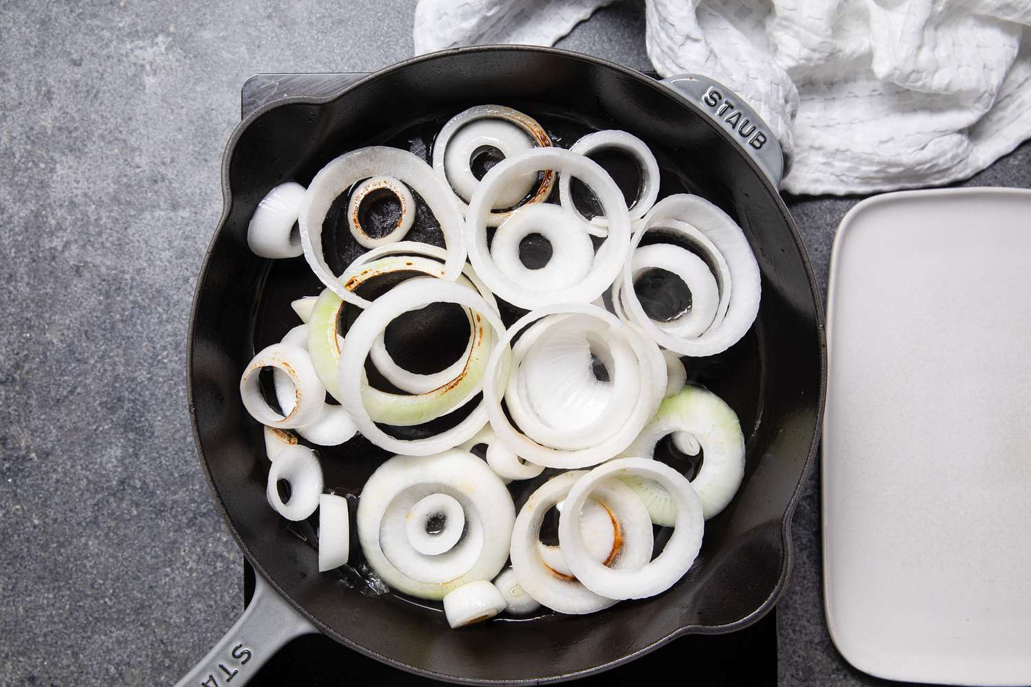 Onions Cooking in a Cast Iron Skillet, and Next to It, an Empty Plate and a White Kitchen Towel
