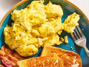 Scrambled eggs served on a plate with toast and bacon a fork placed to the side