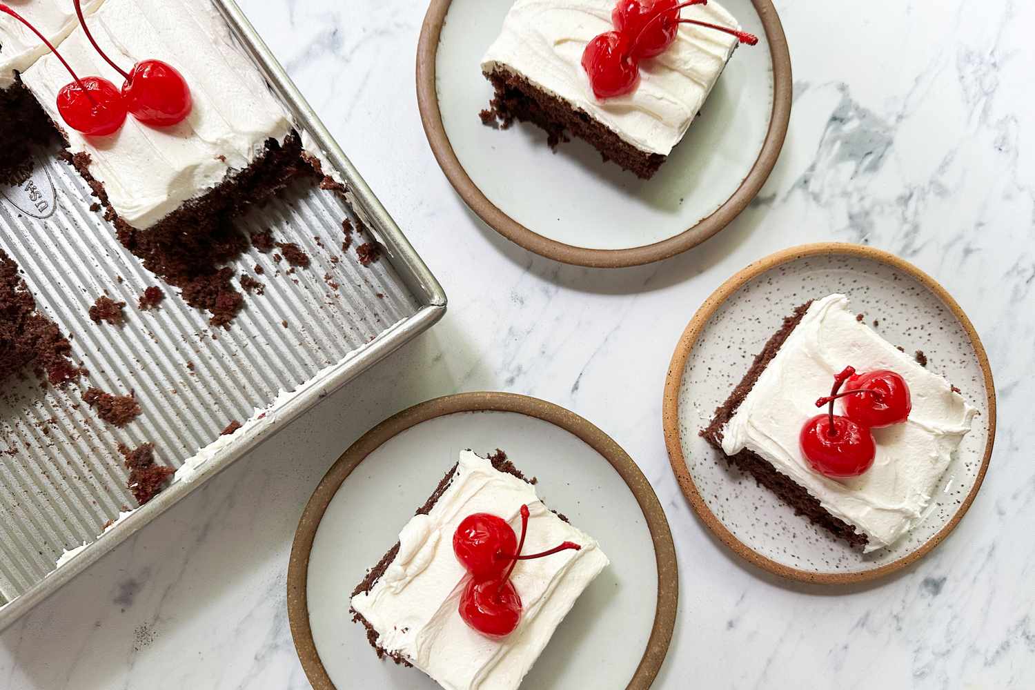 Chocolate cake with white frosting and cherries served on three plates and a baking pan on a marble surface