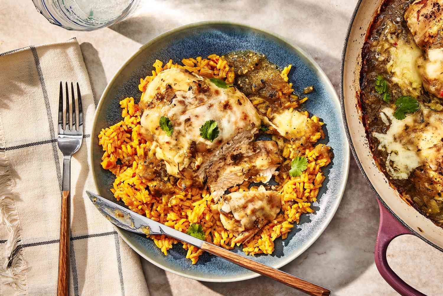 Overhead view of a plate of Salsa Verde Chicken over yellow rice next to a skillet, cloth napkin, fork and drink glass