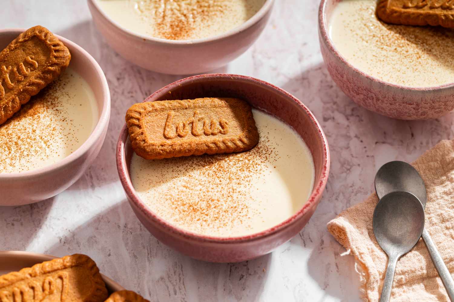 Several bowls of panna cotta each garnished with a Lotus biscuit and dusted with spices two spoons on a napkin nearby