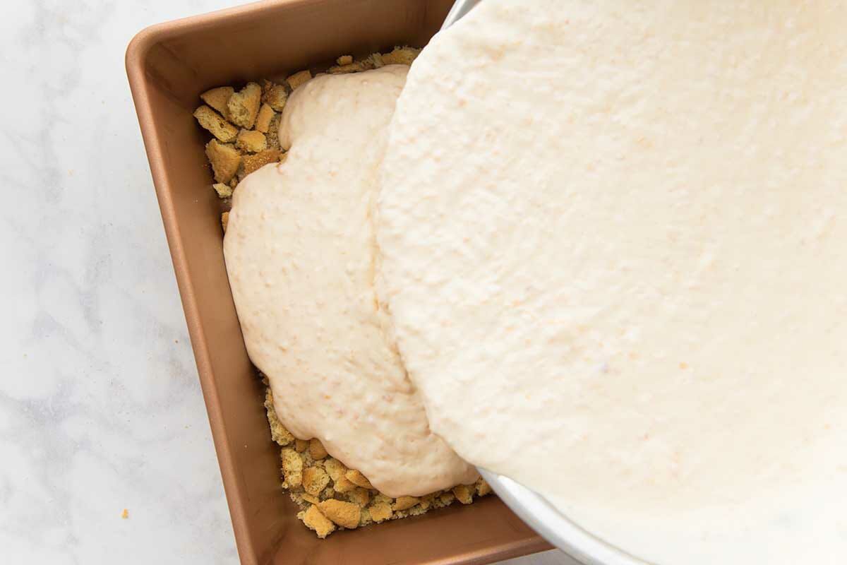 The base of homemade peach cobbler ice cream is being poured over broken vanilla wafers laid in a loaf pan.