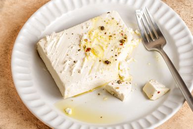 A block of feta cheese garnished with seasoning and olive oil on a white plate with a fork