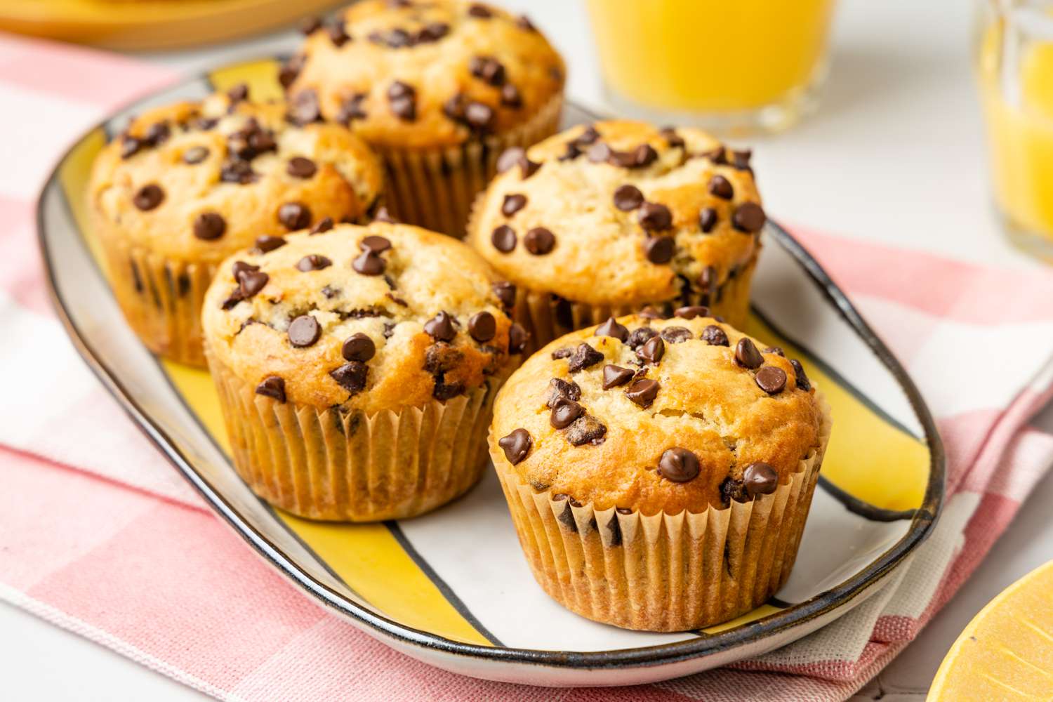 Banana Chocolate Chip Muffins on a Plate Over a Kitchen Towel and Next to Two Glasses of Orange Juice