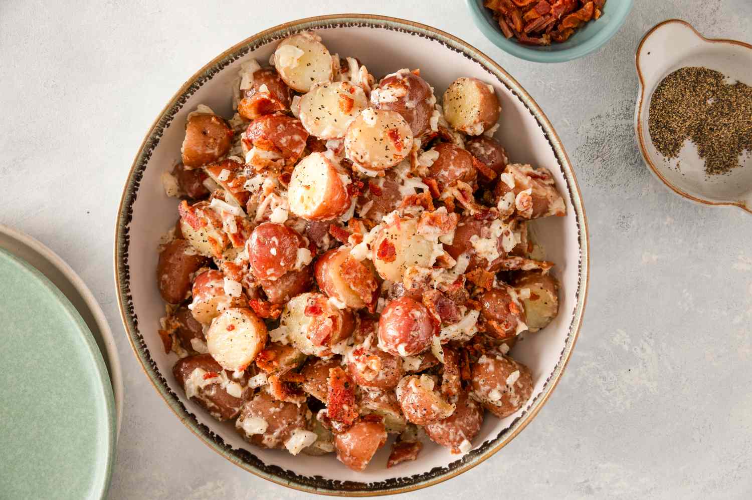 Bowl of bacon potato salad on a table with small bowls of bacon bits and seasoning nearby