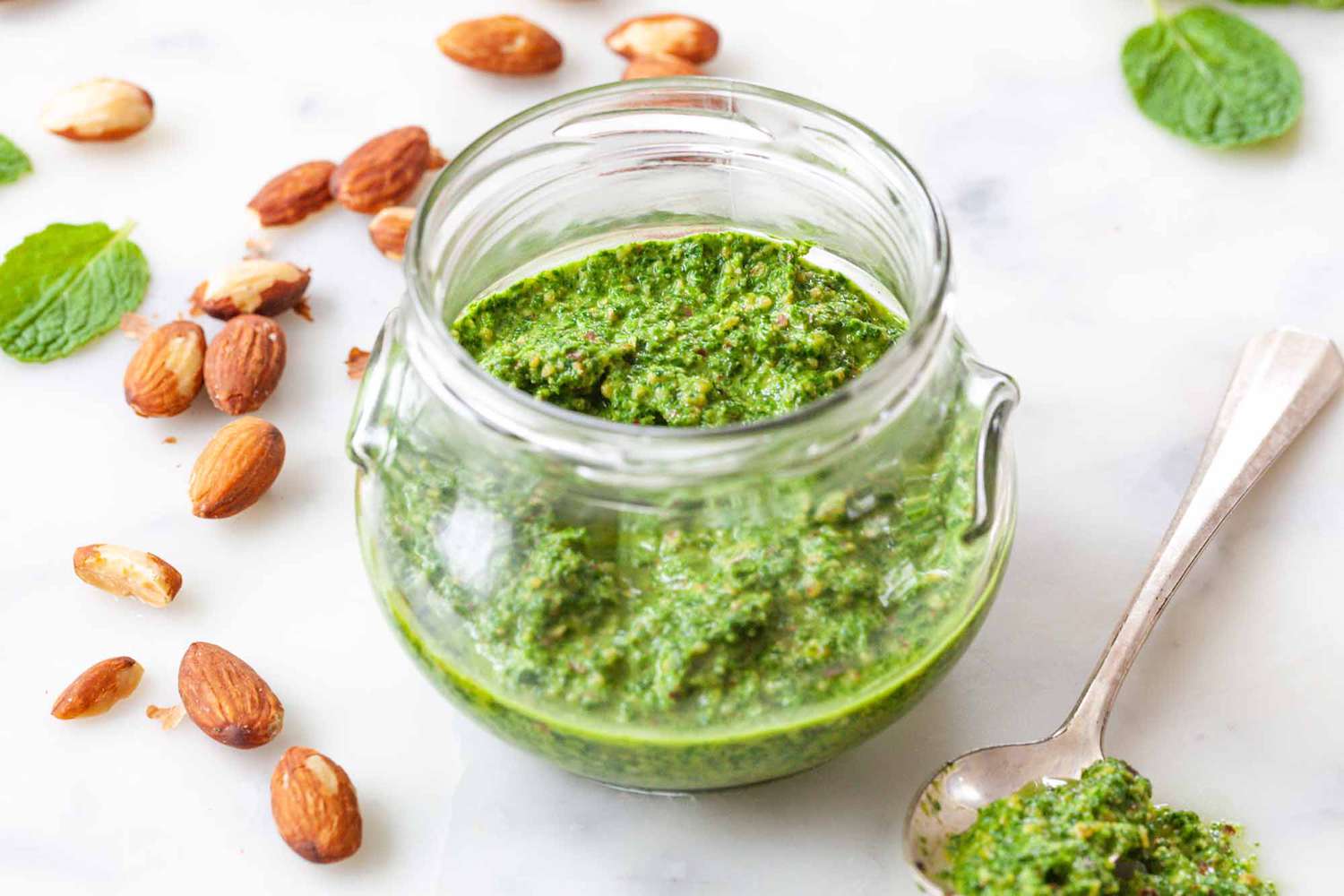 Mint Pesto Sauce - jar of mint pesto on white counter with almonds scattered about and a spoonful of pesto sitting next to the jar