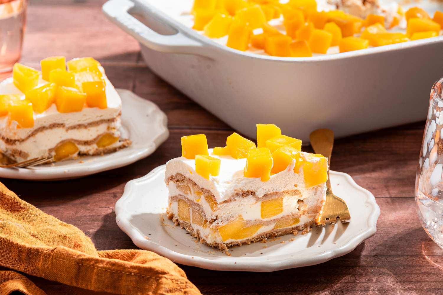 slice of mango royale (mango float) on two plates, and in the background, a dish with more, all at a table setting with glasses and table napkins