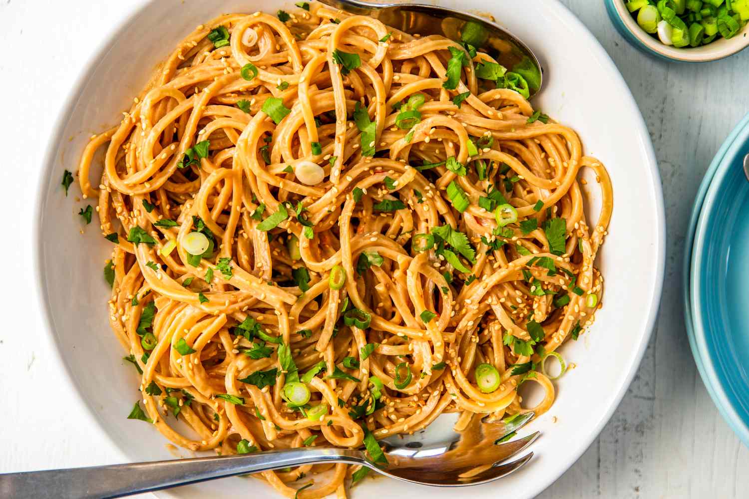 Peanut butter noodles garnished with sesame seeds and scallions in a bowl with serving utensils