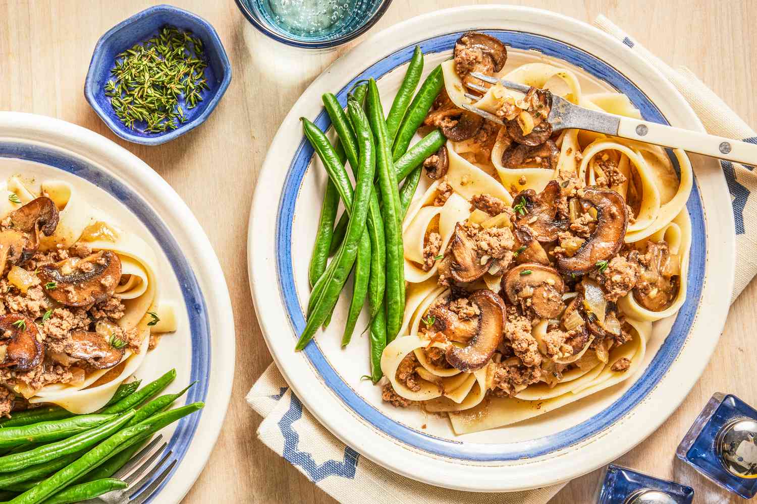 salisbury ground beef with a side of green beans on a plate at a table setting next to another serving on a plate, a small bowl of herbs, and a glass of water