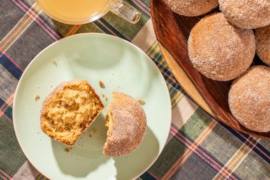 A halved apple cider donut muffin on a plate, and in the surroundings, a cup of apple cider and more muffins stacked on a plate