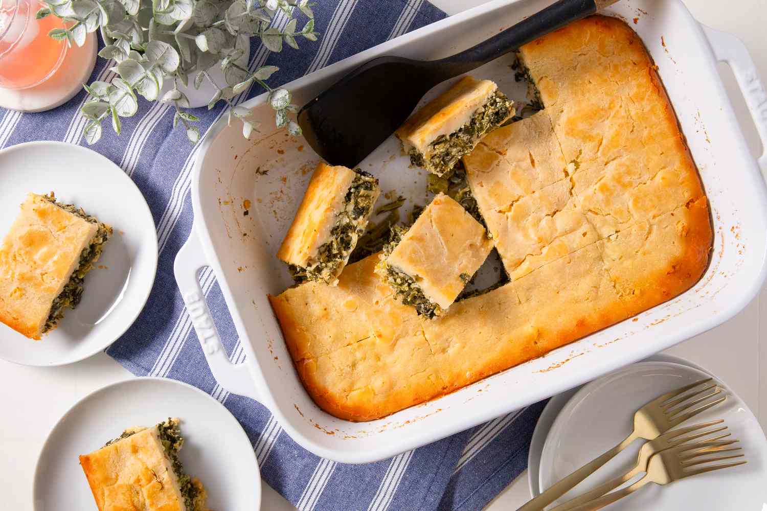 batsaria in a casserole dish and slices of more on small plates at a table setting with a small plant and a stack of plates and utensils