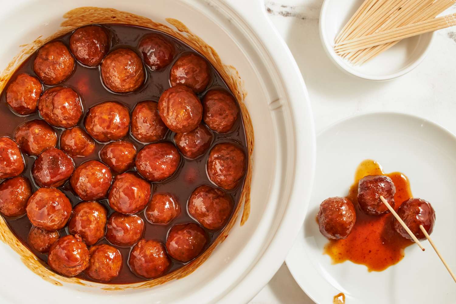 Slow cooker filled with glazed cocktail meatballs with a plate and skewered meatballs to the side