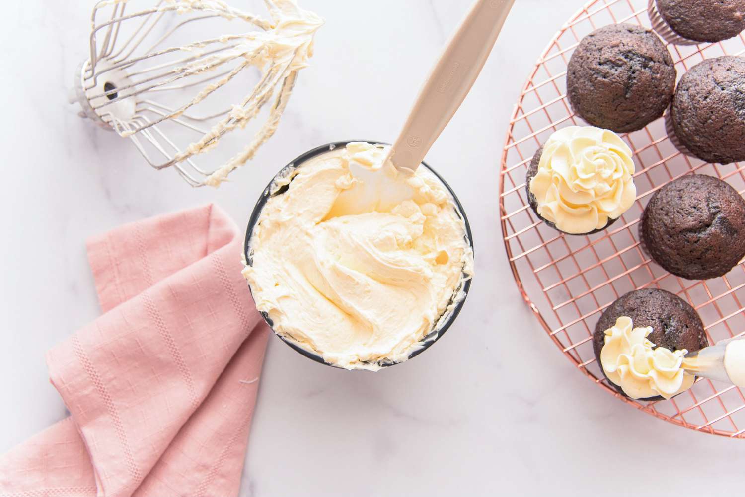 A large bowl of French meringue buttercream next to chocolate cupcakes and a whisk.