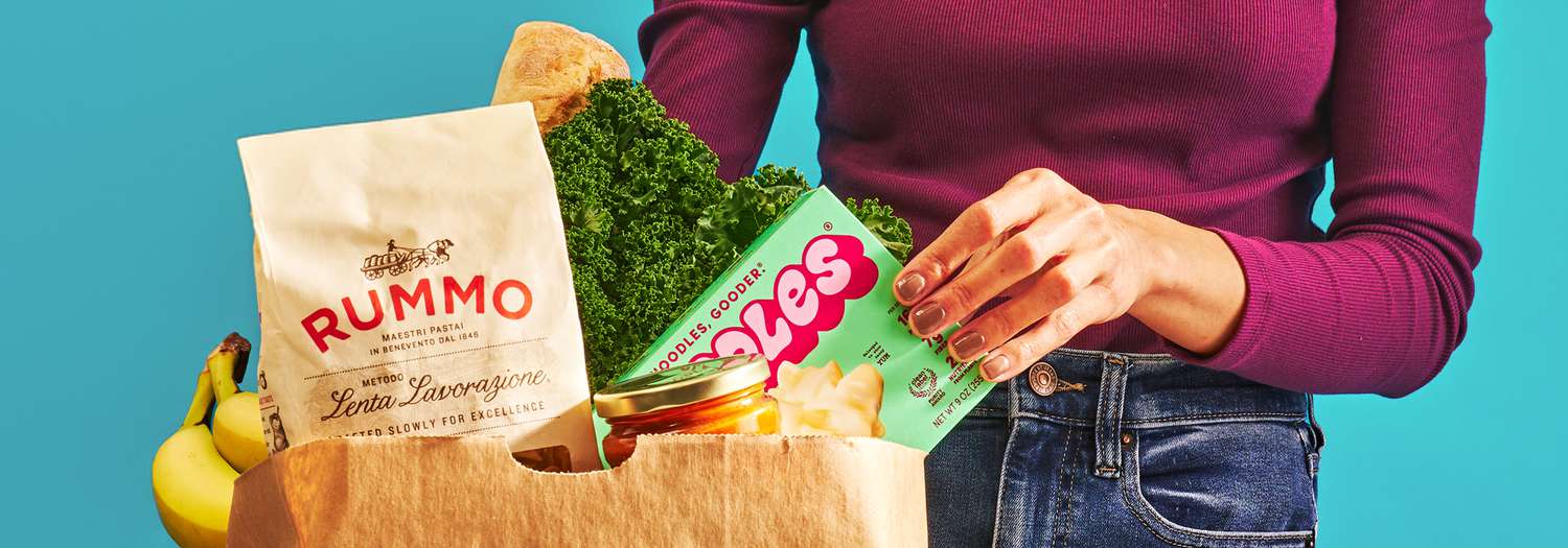 Paper bag of groceries, a woman's hand is pulling out a box of mac and cheese