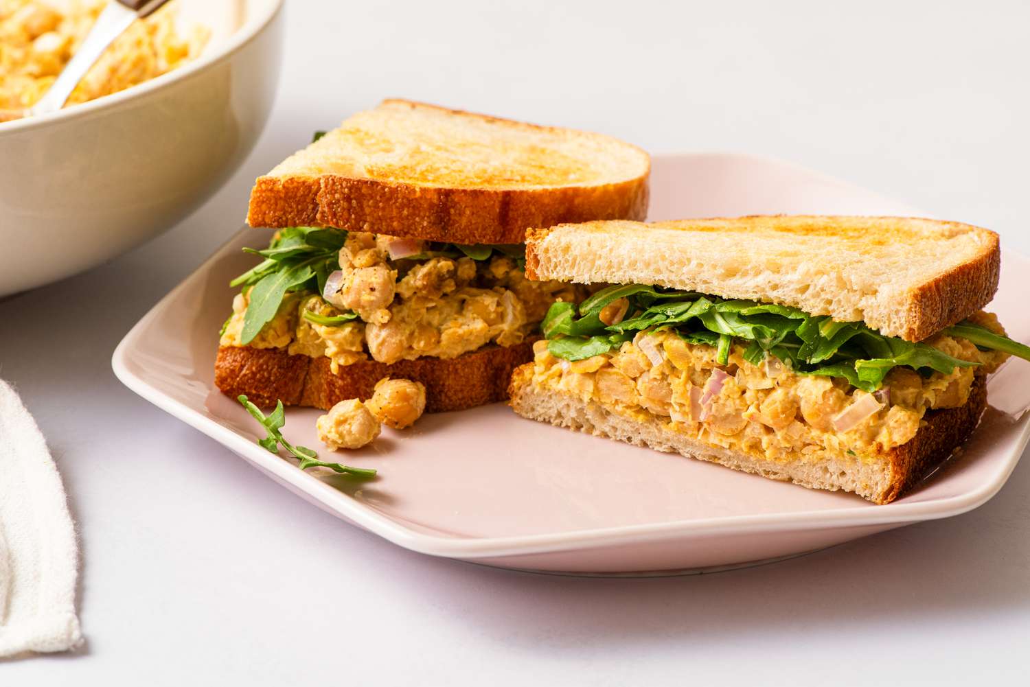 Angled view of a pink plate with two halves of a sandwich made from curried chickpea salad and greens all on a white background
