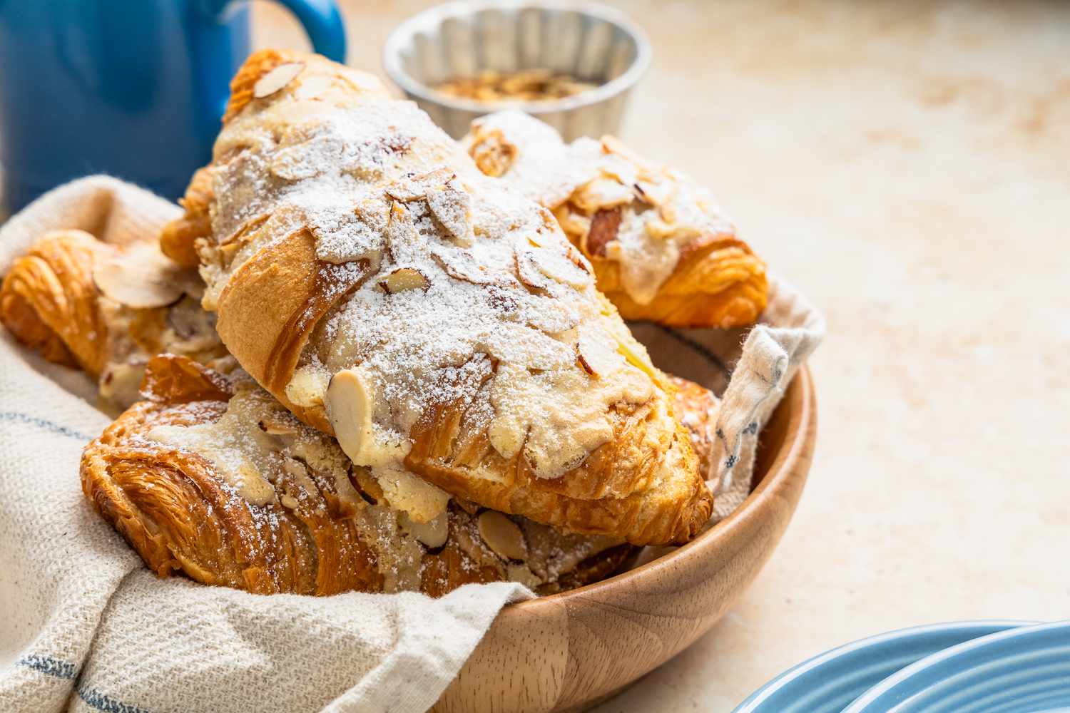 Pile of Almond Croissants on a Wooden Tray, and in the Surroundings, a Kitchen Towel, Bowl of Toasted Almonds, Stack of Light Blue Plates, and a Blue Coffee Mug