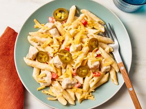 Overhead view of rattlesnake pasta topped cheese and jalapenos light in green plate along with a fork and next to an orange napkin and blue drinking glass on a beige background