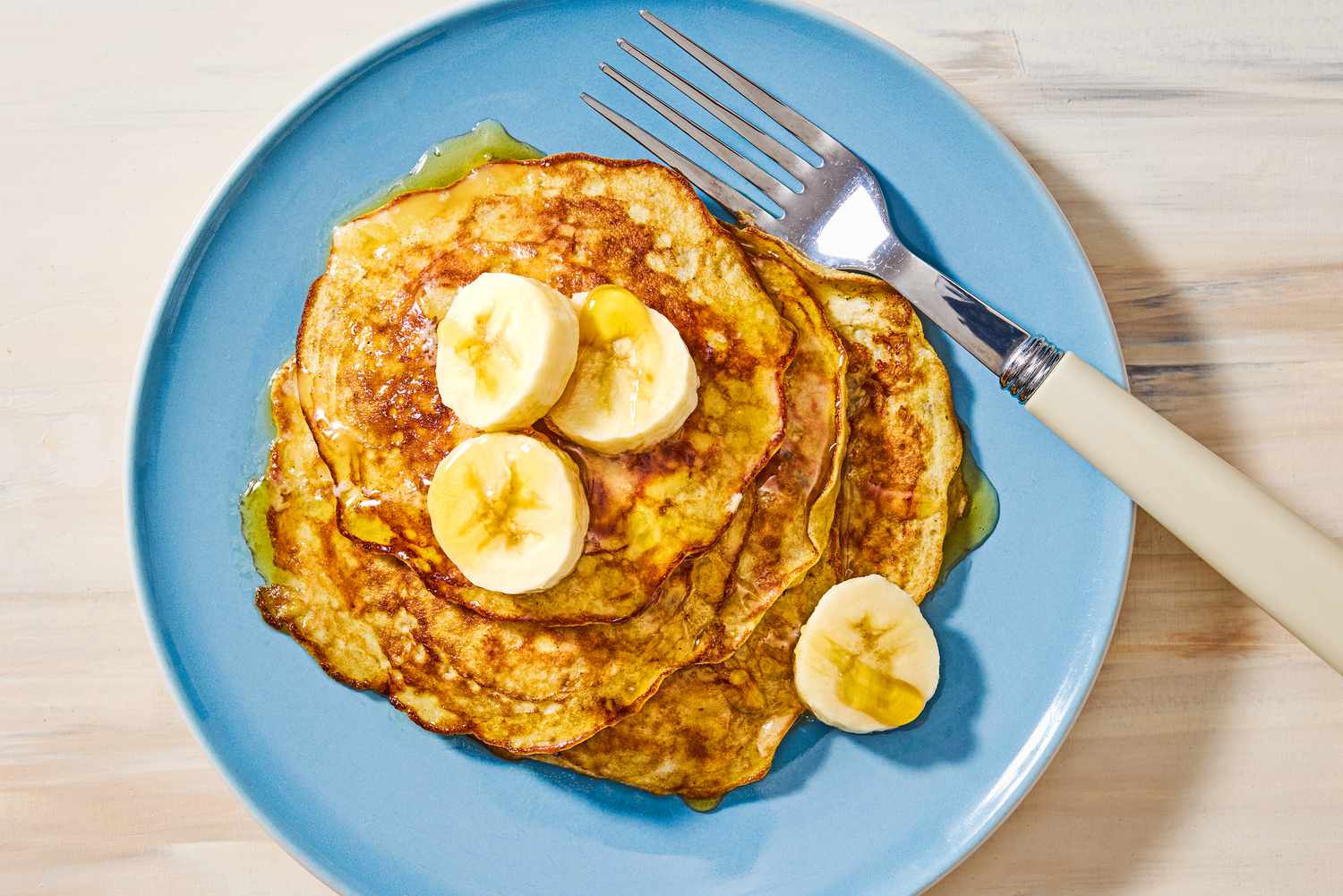 Overhead view of a blue plate of 2-ingredient pancakes topped with bananas next to a fork, all on a wooden tabletop