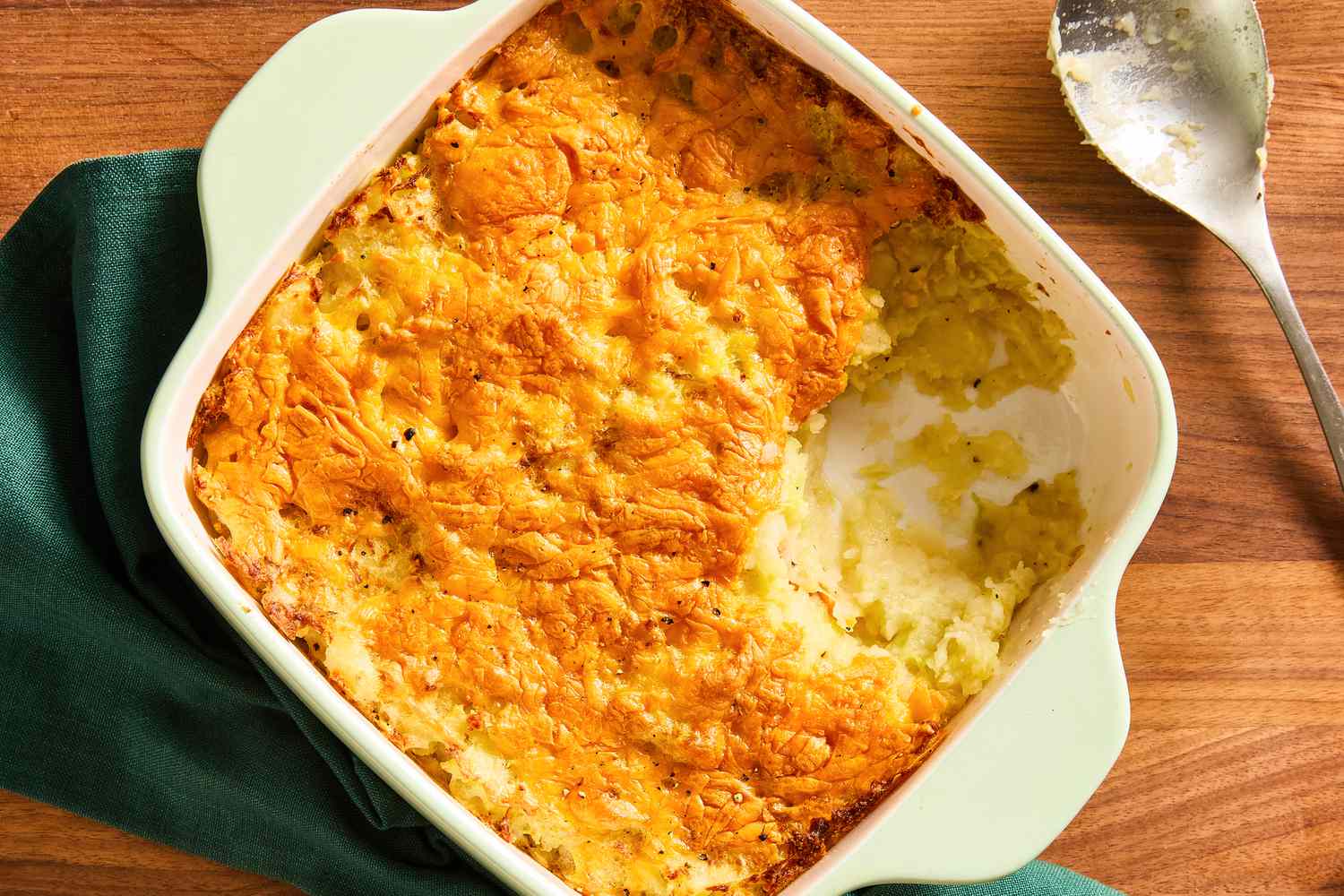 Overhead view of a square white baking dish of potato cabbage casserole sitting on a dark green napkin next to a serving spoon all on a wooden tabletop