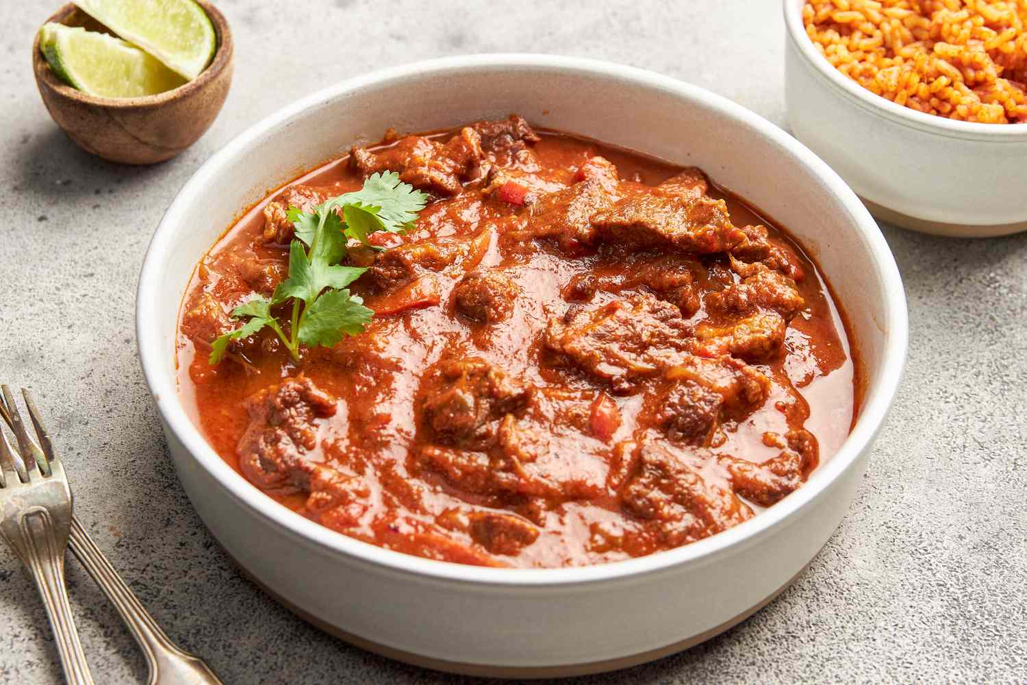 Bowl of carne picada with a sprig of cilantro and in the surroundings, a bowl of seasoned rice, a small bowl of sliced limes, and utensils (on the counter)
