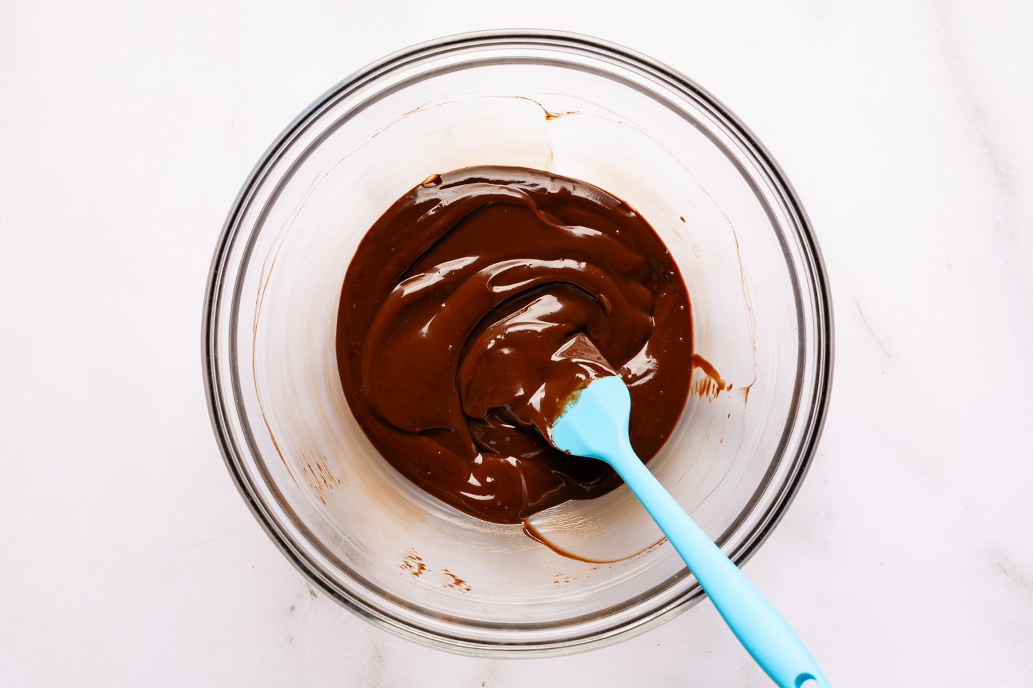 Overhead view of a clear glass bowl with melted cream and chocolate mixture along with a blue spatula for Easy Brooklyn Blackout Cake recipe