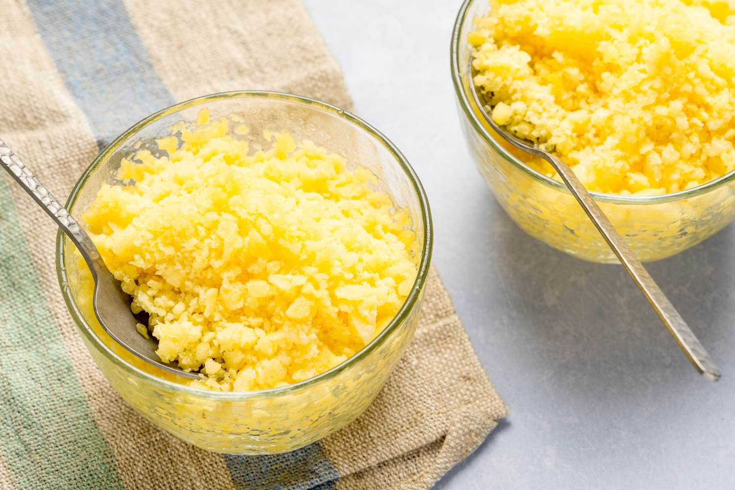 Two bowls of pineapple coconut granita served with spoons placed on a textured cloth