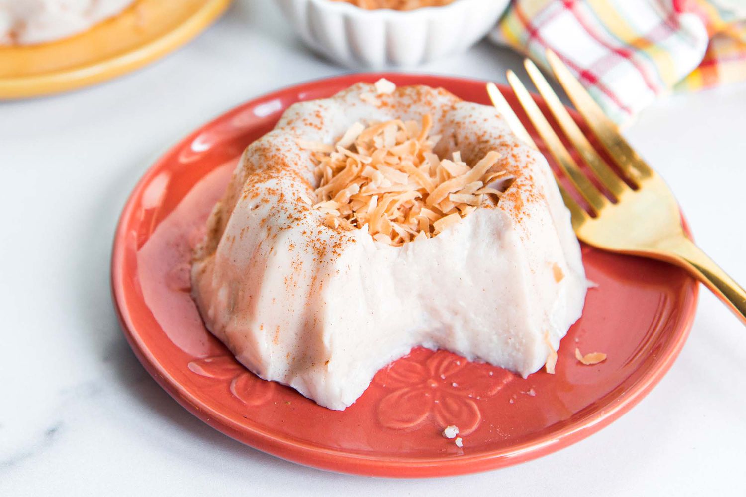 Homemade bundt shaped templeque is on a coral plate with a gold spoon set on the left side. The tembleque has toasted, shredded coconut mounded in the center and a section of the pudding is missing. Behind the plate is a striped dish towel, small white container and yellowl plate all in partial view.