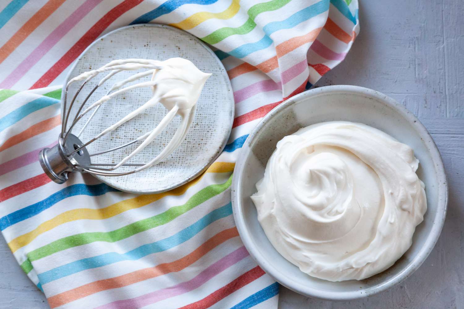 Whipped cream frosting with cream cheese in a bowl with a whisk next to it and a striped linen underneath.