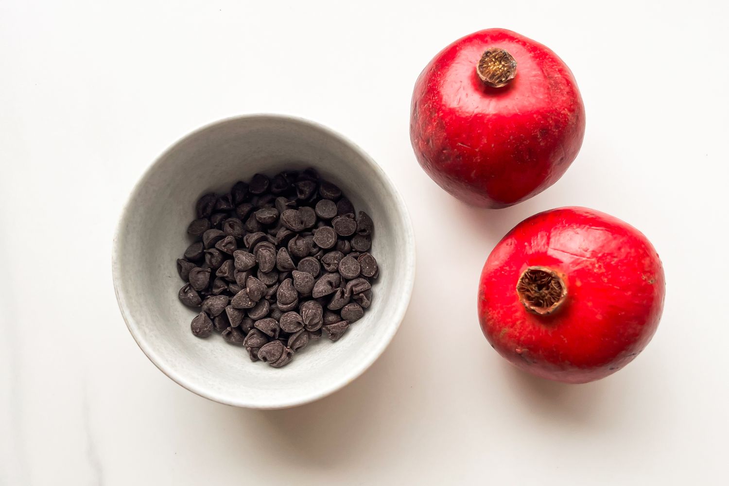 A bowl of chocolate chips and two pomegranates on a white surface