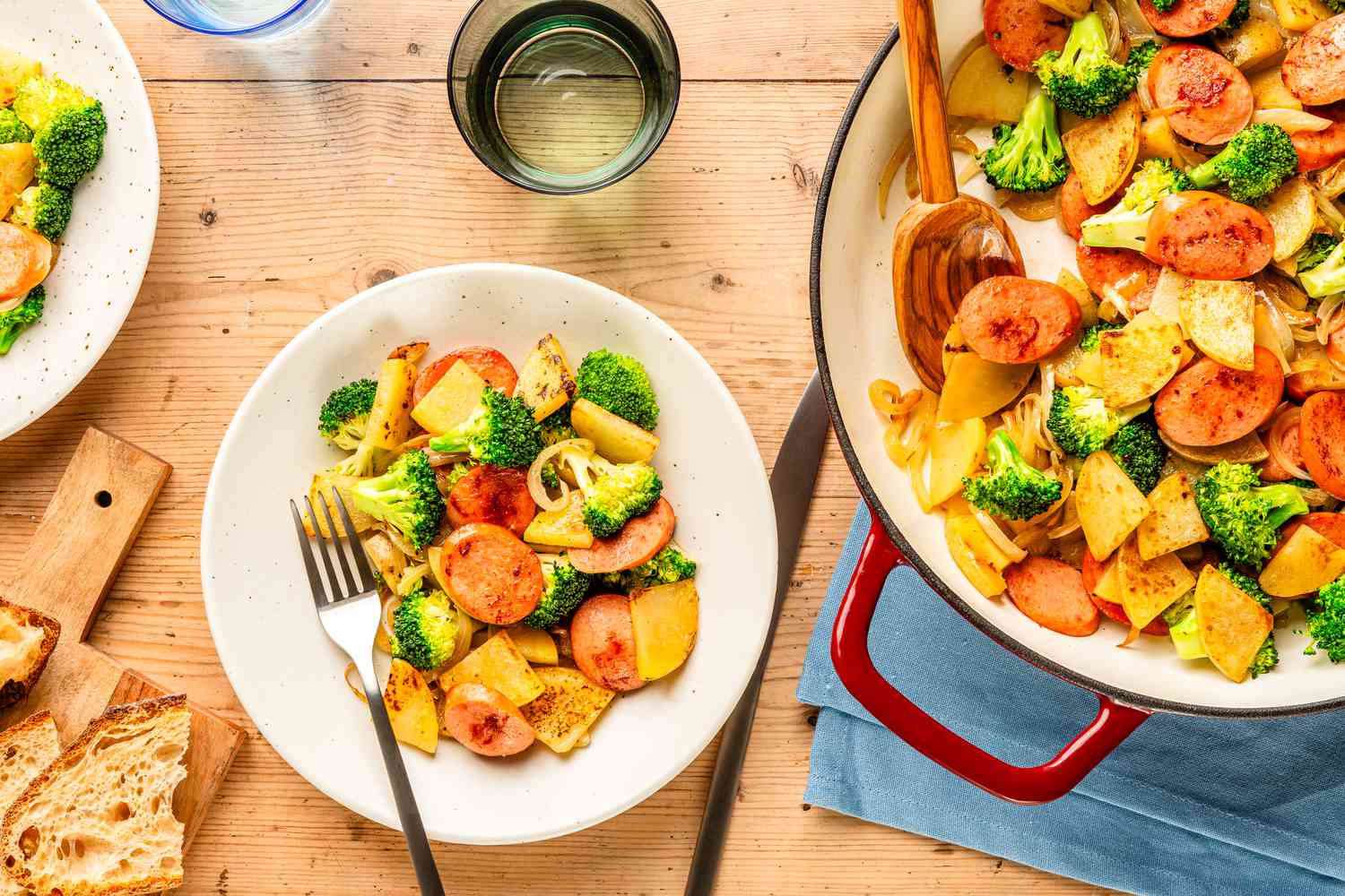 Overhead view of a white bowl of Kielbasa, Potato, and Broccoli Skillet with a fork next to a skillet, bread on a cutting board and a drinking glass 