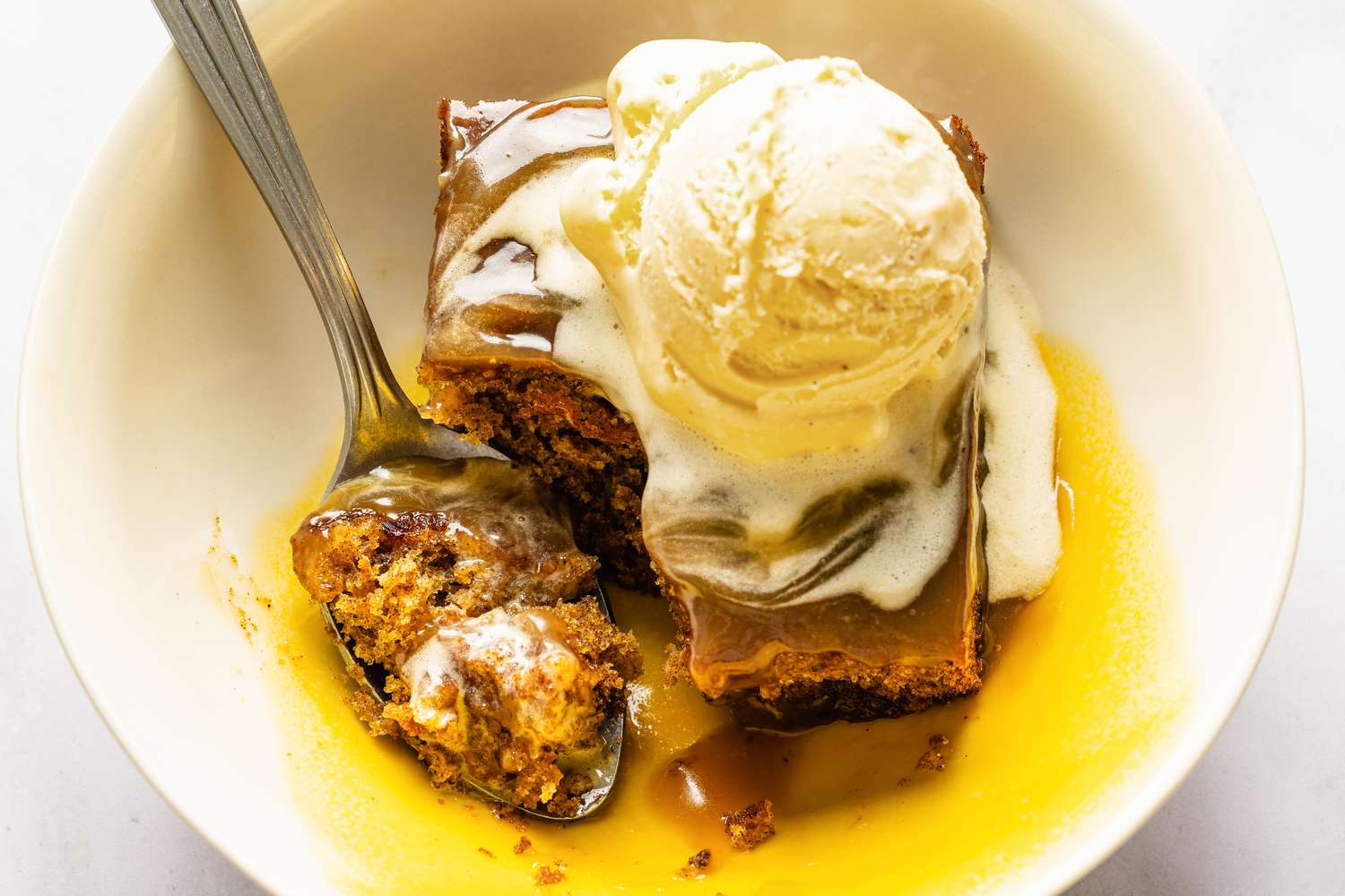 A bowl containing sticky toffee pudding topped with a scoop of vanilla ice cream and a spoon resting in the dish