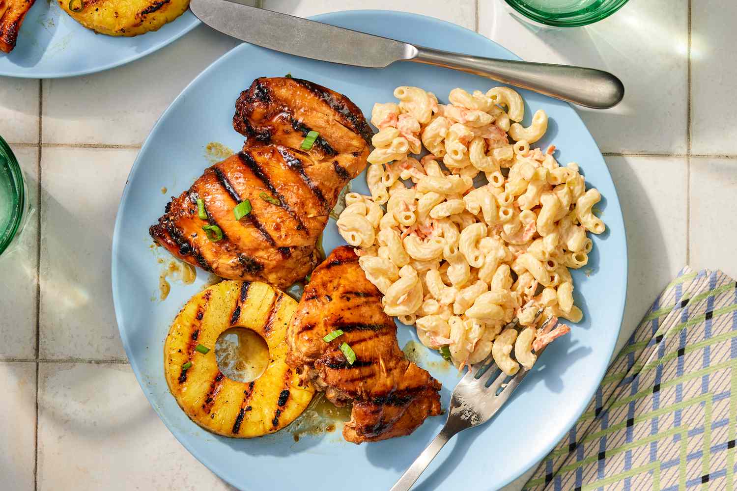 Plate with grilled chicken, pineapple, and macaroni salad, placed on a tabletop with cutlery and glasses nearby