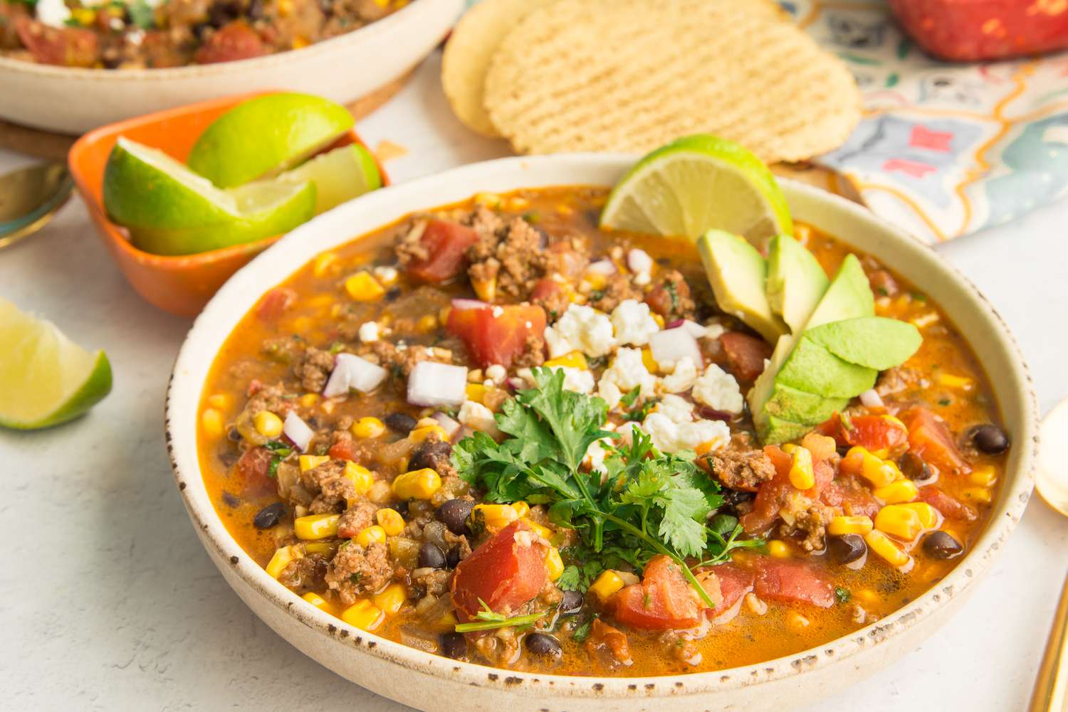 Bowl of Stovetop Taco Soup Topped with Avocado Slices, Cotija Cheese, and Cilantro and Surrounded by Bowls of Toppings