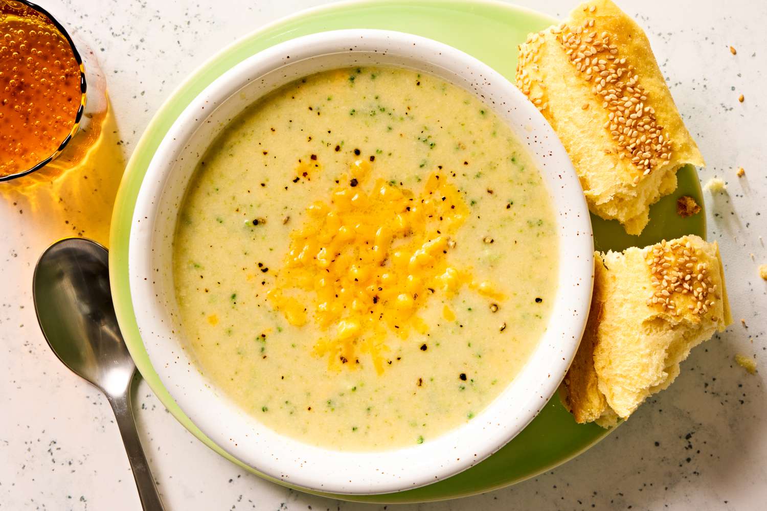 Overhead view of a bowl of broccoli cheddar soup on a plate with pieces of bread and next to a spoon and drink glass