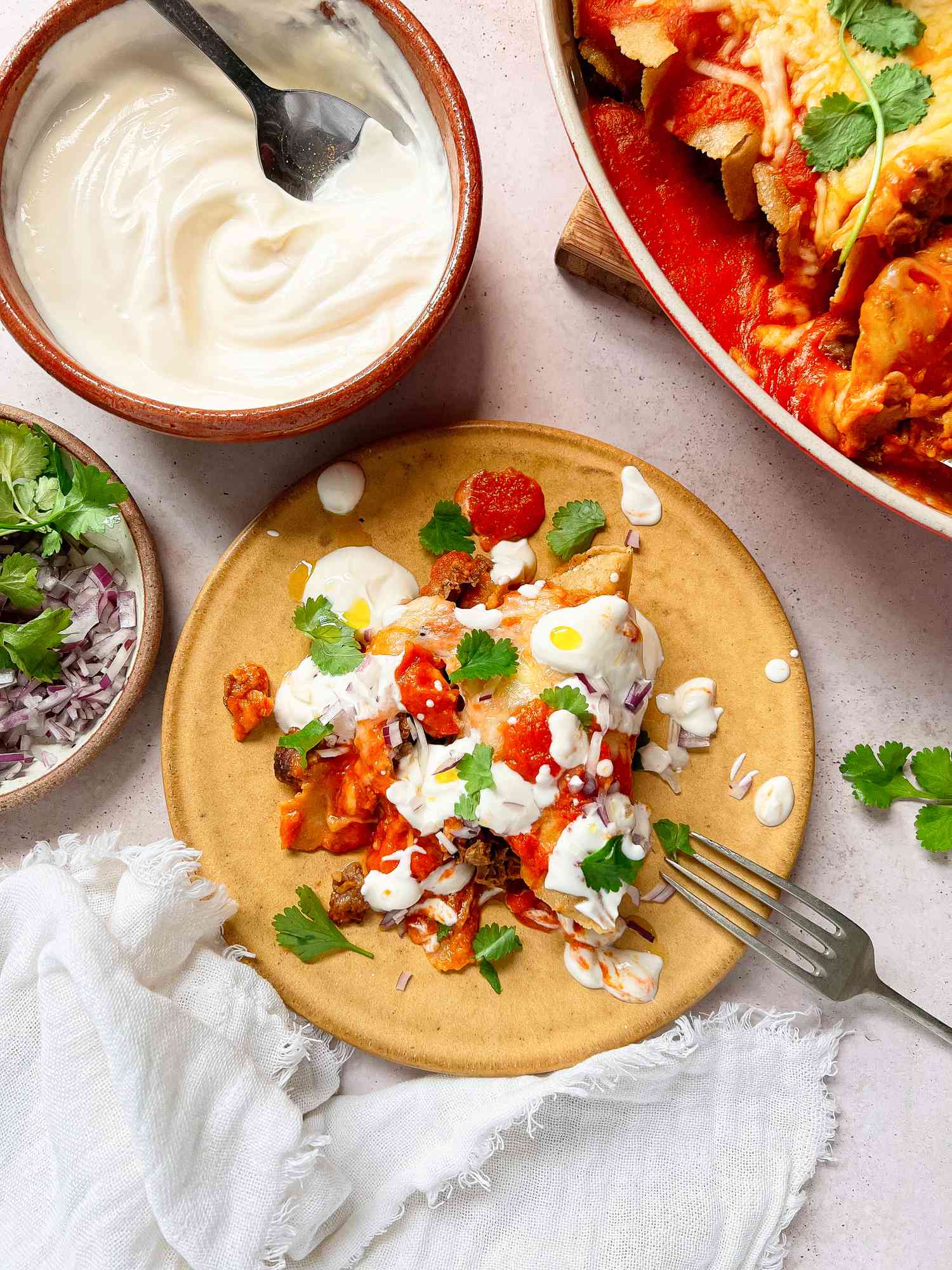 Pulled Pork Enchiladas on a Plate Topped with Cilantro, Sour Cream, and Onions, Surrounded by a Plates and Bowls of Toppings and the Casserole Dish with More Enchiladas
