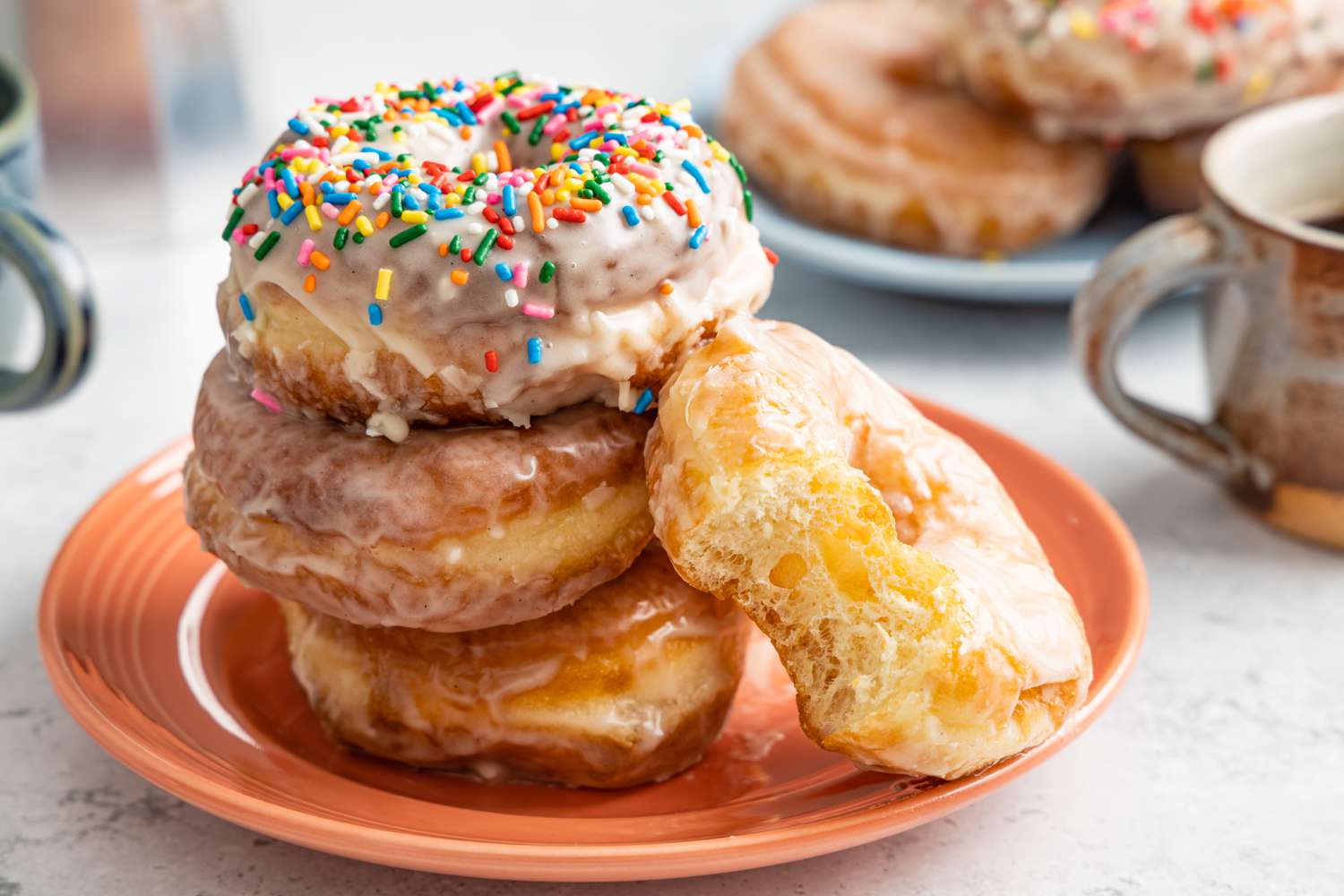 Stack of Glazed Donuts with the Top Donut Covered in Sprinkles, and Leaning Next to the Stack, a Glazed Donut with a Bite Missing, All on a Platter. In the Background, a Two Mugs of Coffee and Another Platter of Glazed Donuts