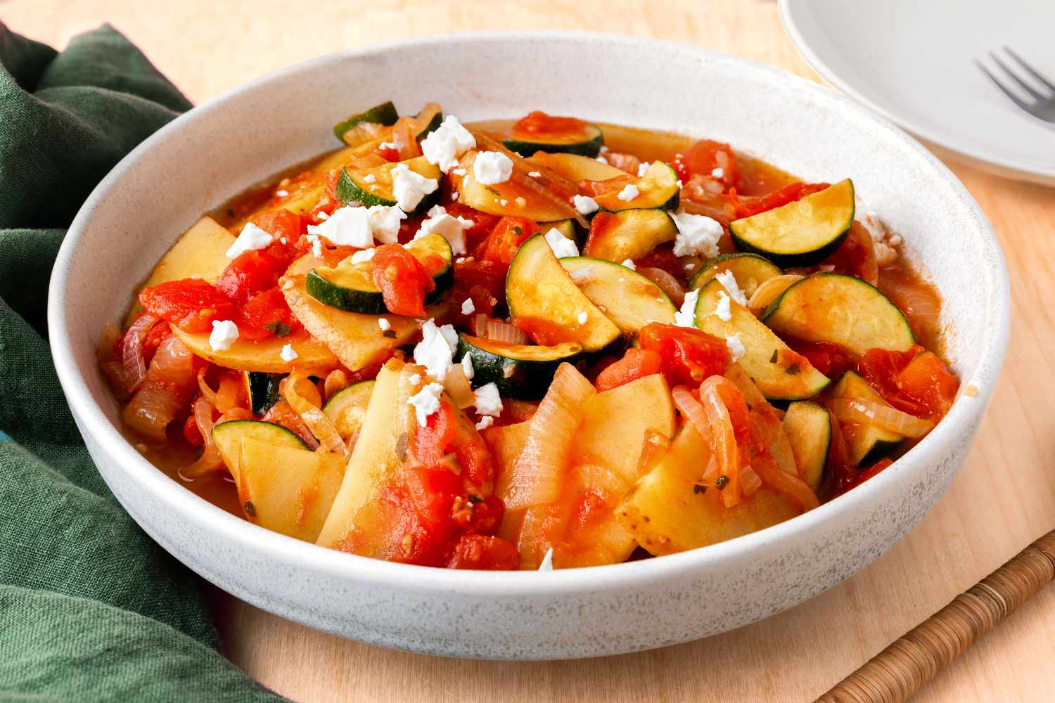An angled view of a white bowl of Zucchini Con Patate on a table next to a spoon and a green napkin