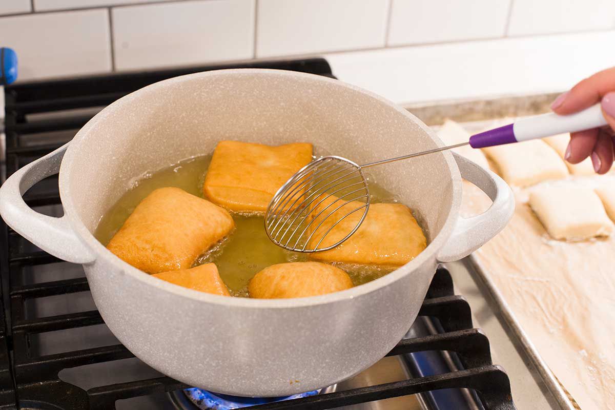 New Orleans Beignets are in a dutch oven on the stove being turned in the hot oil. One side is golden brown. A slotted spoon is being used to turn them.
