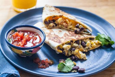 Side view of two simple breakfast quesadillas on a blue plate with a bowl of salsa to the left and a glass of orange juice above. The scrambled eggs, black beans and cilantro are visible inside the quesadilla.