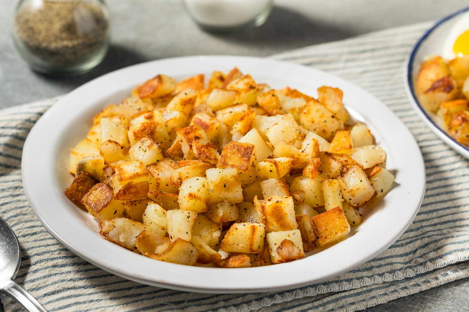 Angled view of a white bowl of breakfast potatoes in a table setting with a spoon and salt and pepper shakers in the background