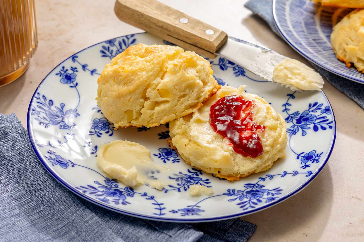 Angled view of a decorative small plate with one sliced mashed potato biscuit with butter and jam next to a butter knife with butter on the end