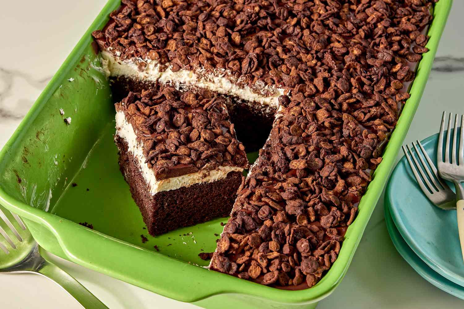 A rectangular dessert in a green baking dish topped with chocolate chips and missing a square piece showing layers of cake and cream filling
