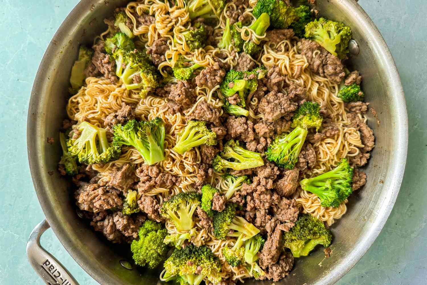 Overhead view of a pot of ground beef, broccoli and noodles on a light blue background