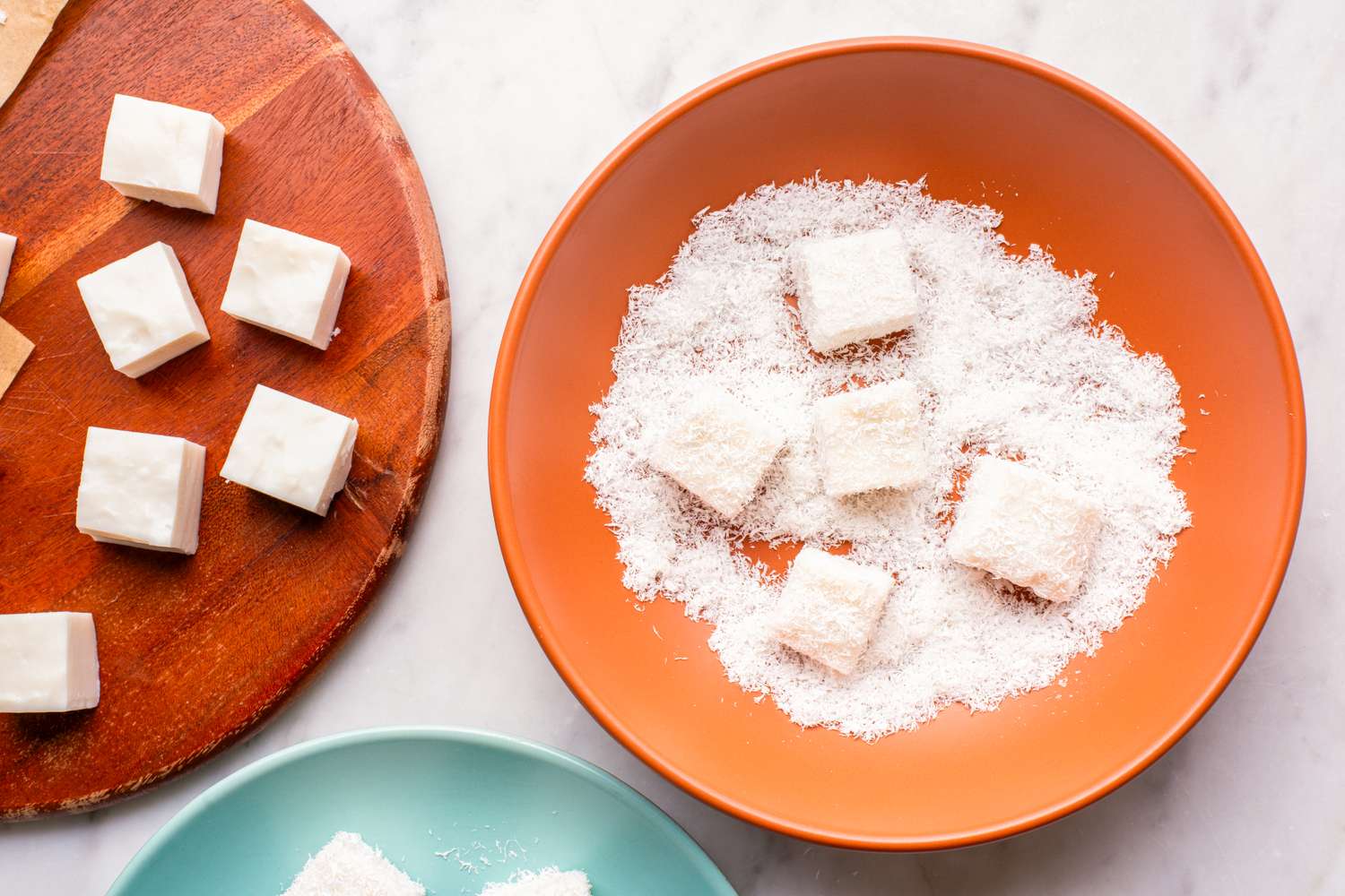 Coconut milk pudding squares being dipped into shredded coconut in an orange bowl, with prepared and uncoated squares visible